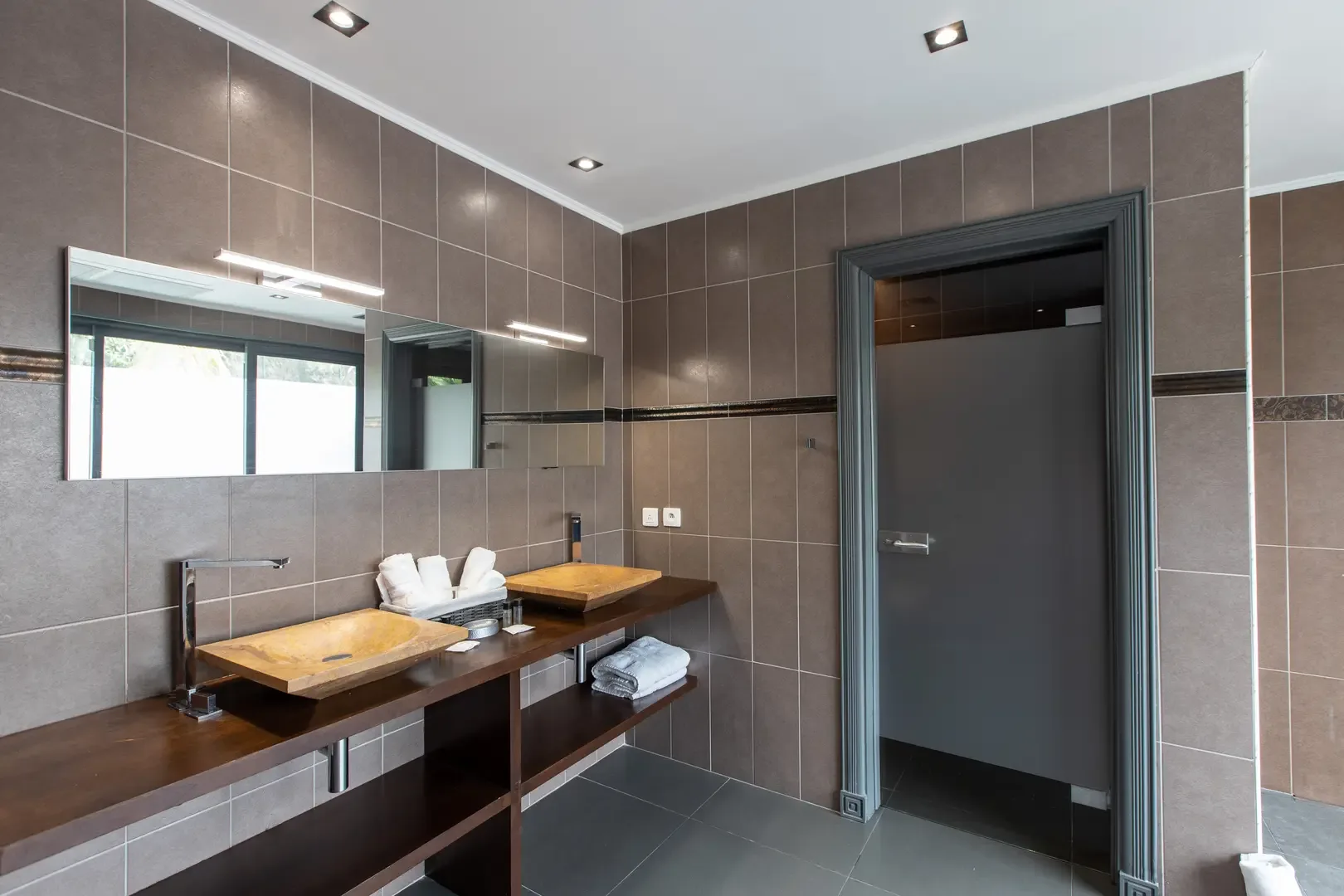 Stone vessel sinks on a dark wood floating shelf complement the large horizontal mirror with integrated lighting and warm brown tiled walls.