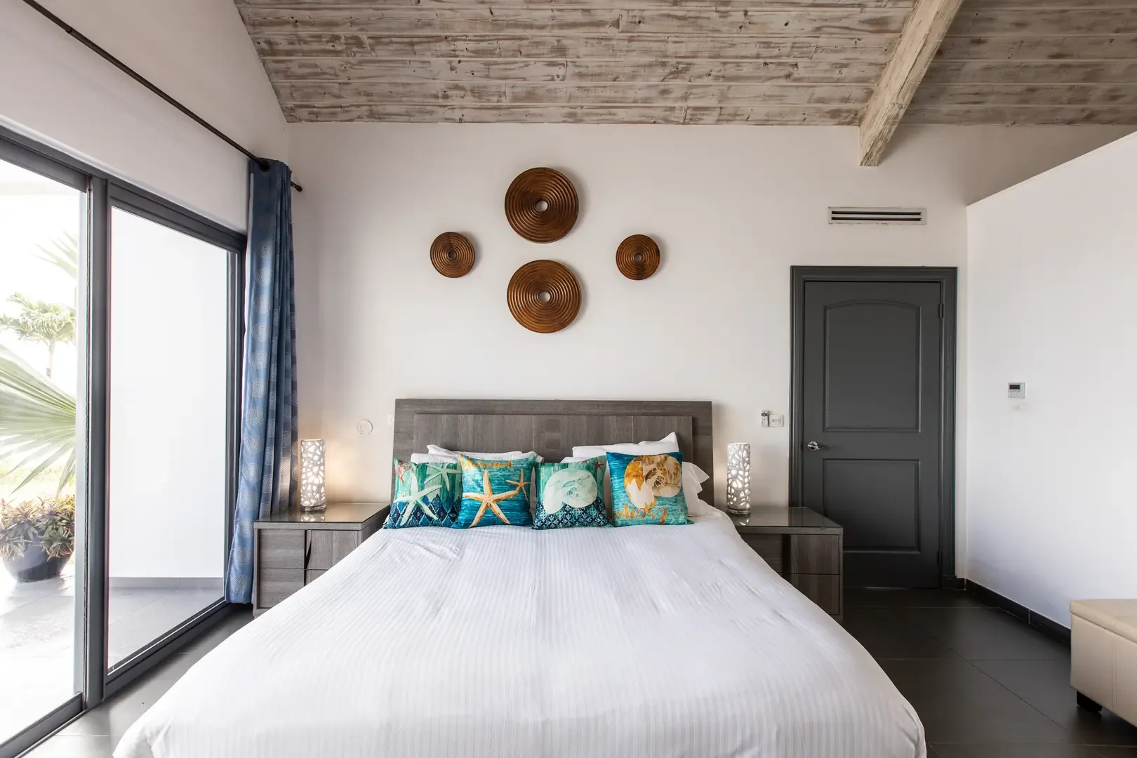 Wake up to paradise in this bright bedroom featuring beach-themed pillows, a grey wooden headboard, and sliding glass doors that invite the Caribbean breeze.