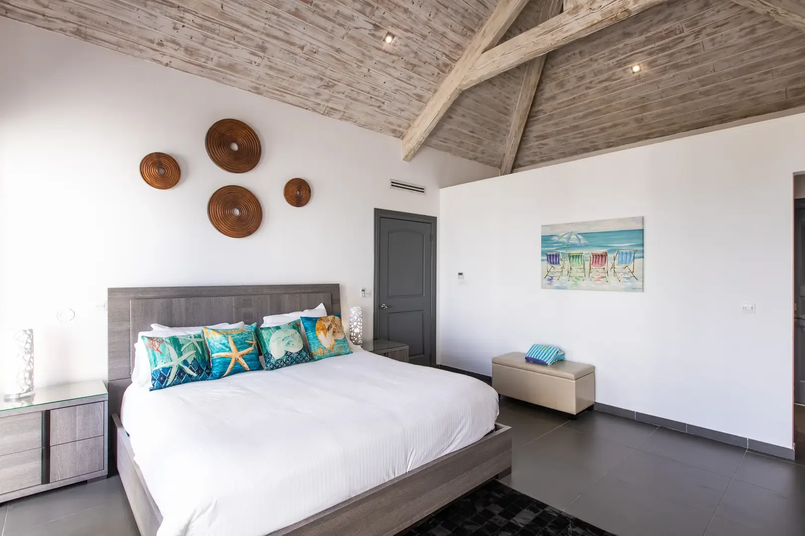 Beach-inspired pillows and artwork complement the high vaulted wood-beamed ceilings, while four circular wall decorations add artistic flair to this coastal-themed bedroom.