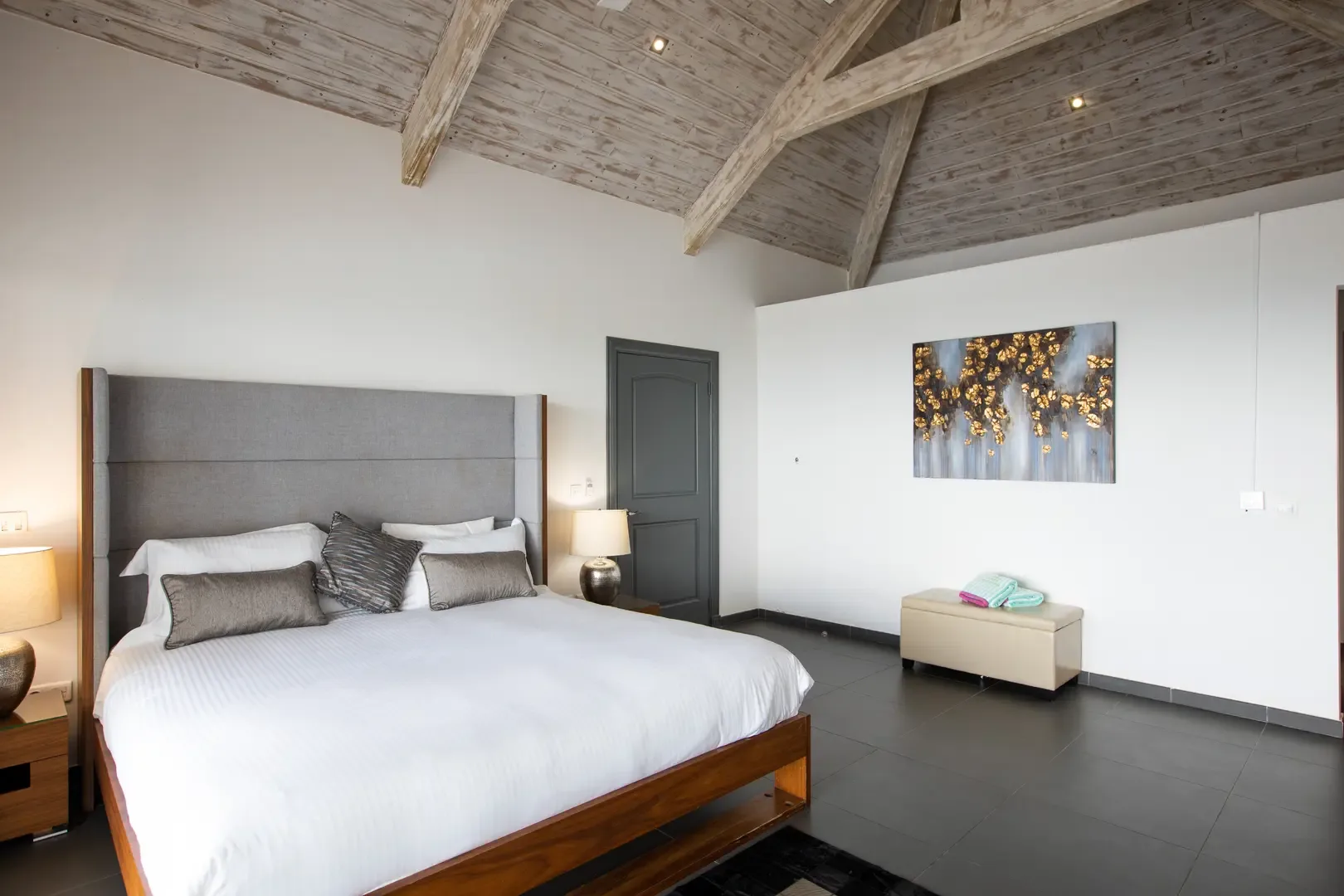 High ceilings with exposed wood beams frame the tall grey headboard, while neutral decor and abstract wall art create an elegant, serene atmosphere.