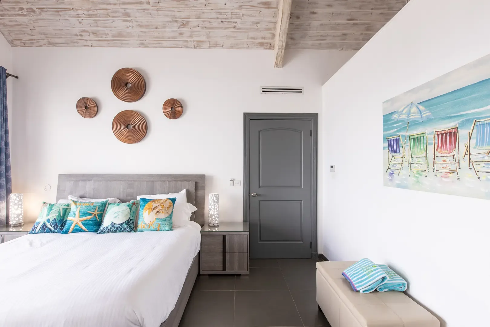 Coastal elegance defines this bedroom with nautical-themed pillows, lantern lamps on matching nightstands, and circular spiral wall art beneath high white-washed wood ceilings.