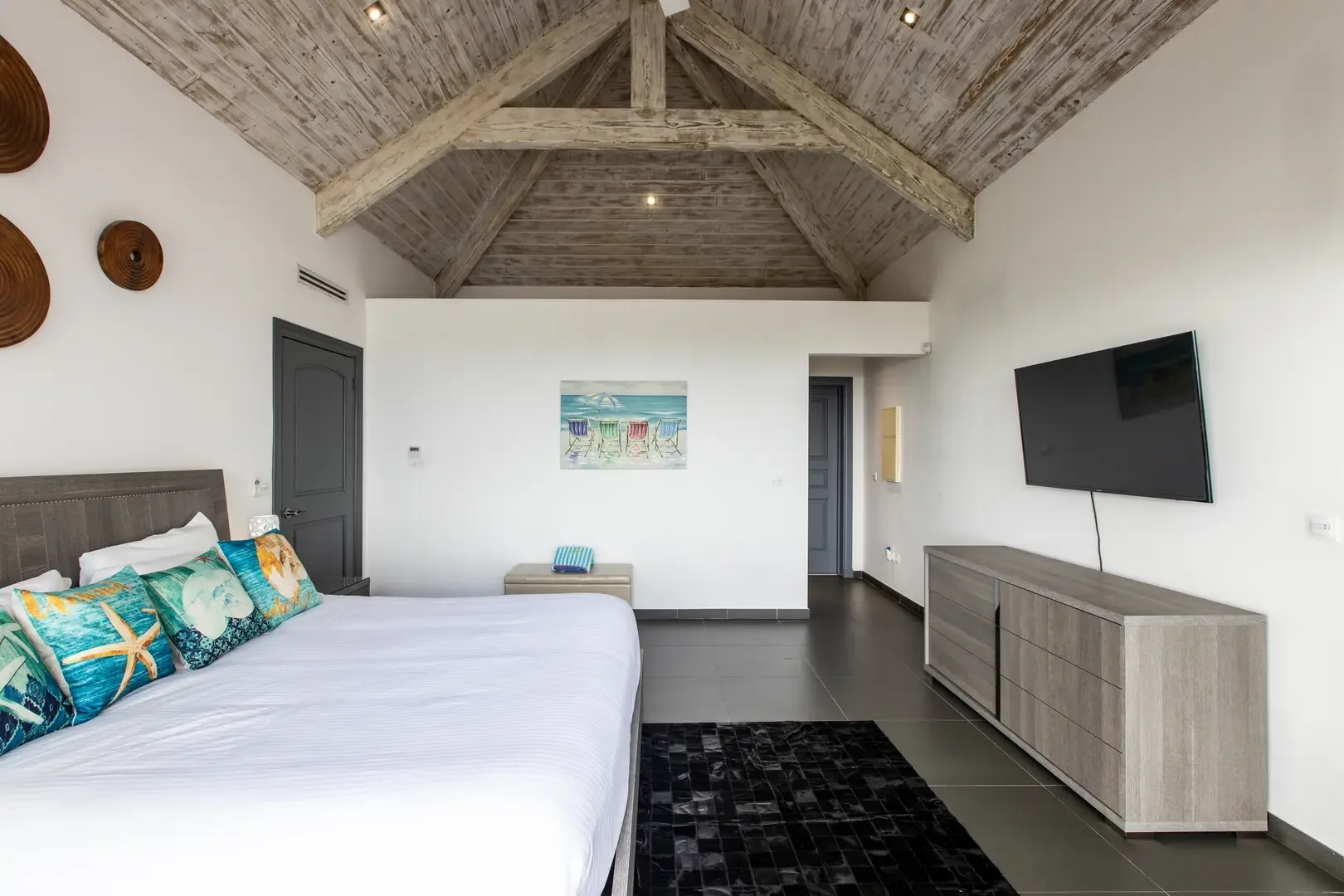 A spacious bedroom with vaulted weathered-wood ceiling, beach-themed pillows, flat-screen TV over a wooden dresser, and beach-inspired wall art creates a relaxing retreat.