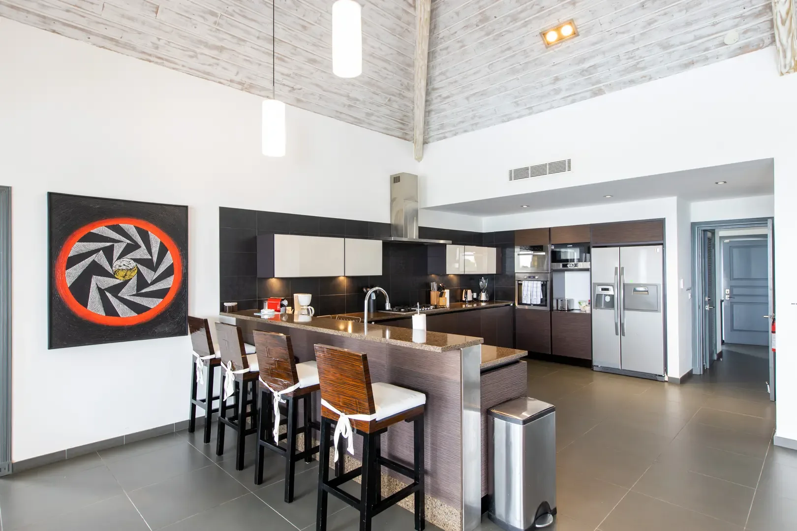 The gourmet kitchen features a breakfast bar with four wooden stools, stainless steel appliances, and vaulted whitewashed ceilings—perfect for morning coffee or evening cocktails.