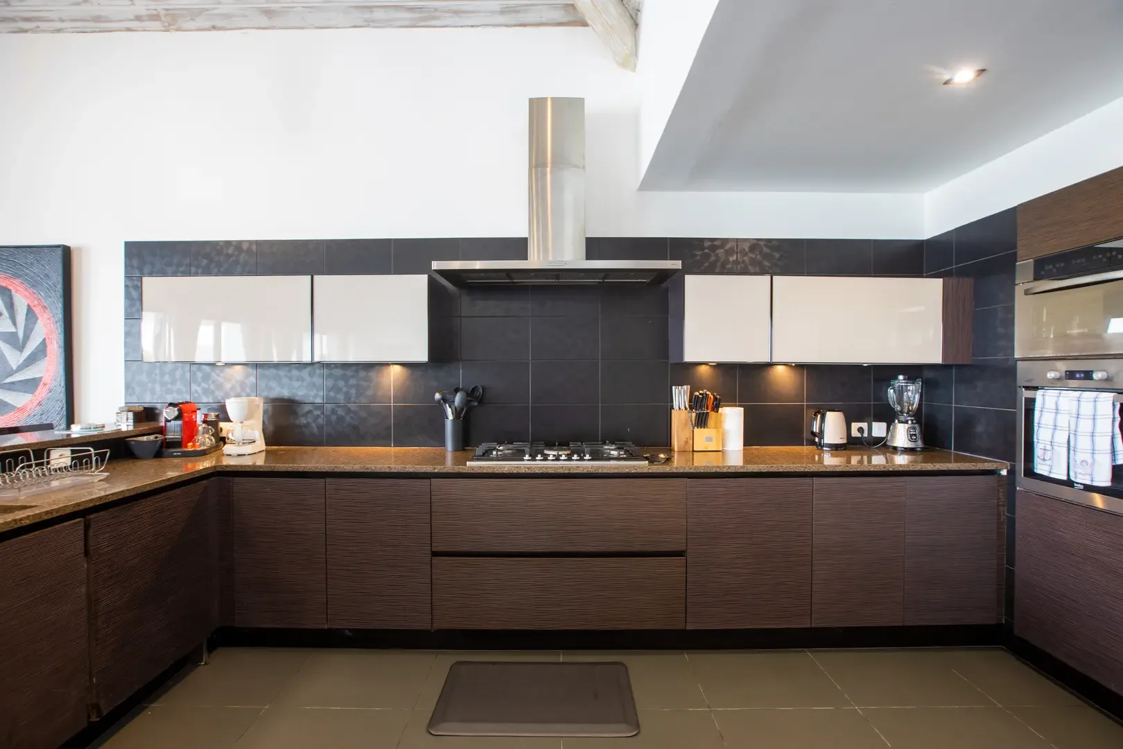 Dark textured cabinetry contrasts with white minimalist upper cabinets and under-lighting, while integrated appliances and a dark tiled backsplash create a sophisticated culinary space.