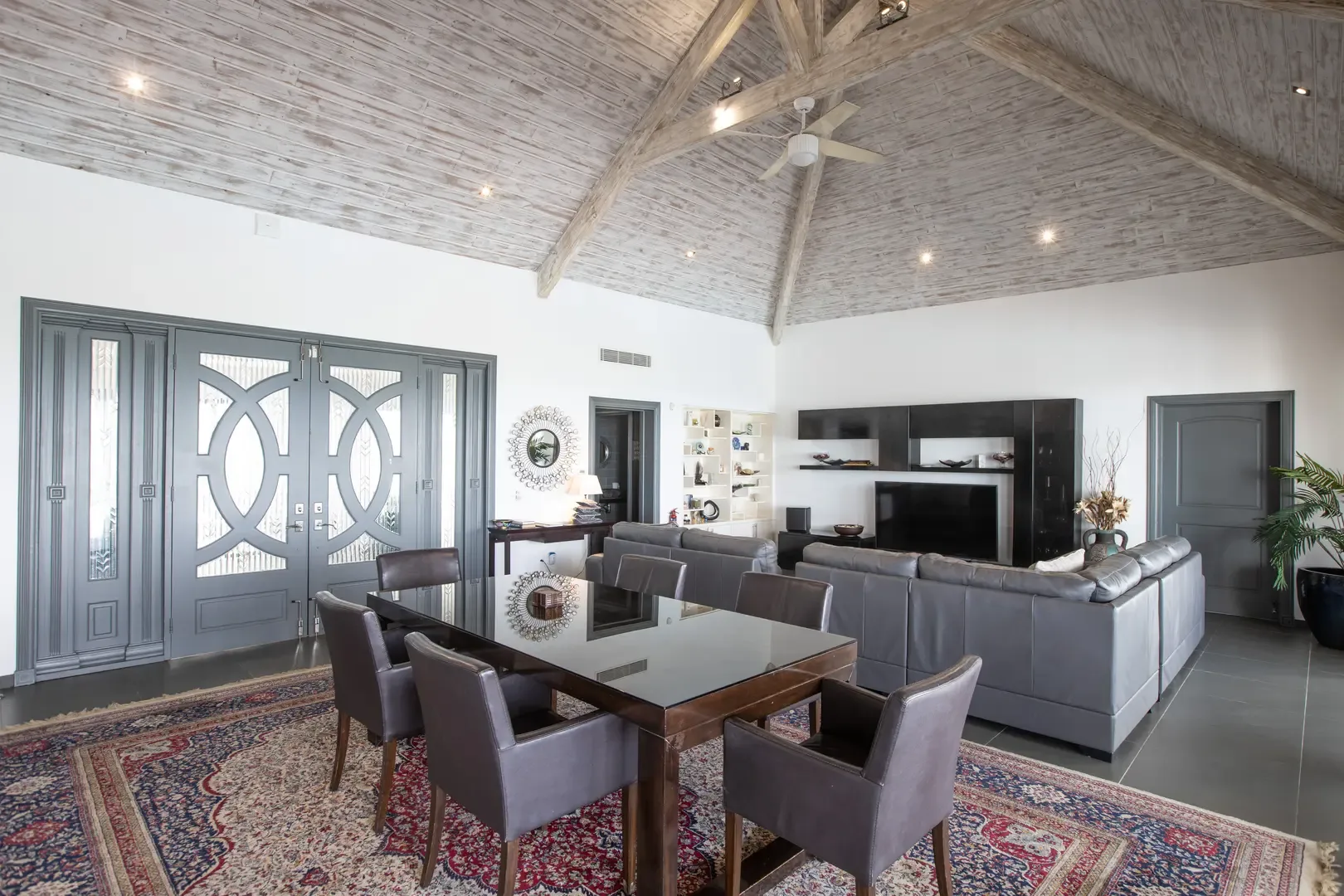 The open-plan living and dining area showcases vaulted wood-planked ceilings with exposed beams, a gray leather sectional, and dramatic dark gray architectural doors.