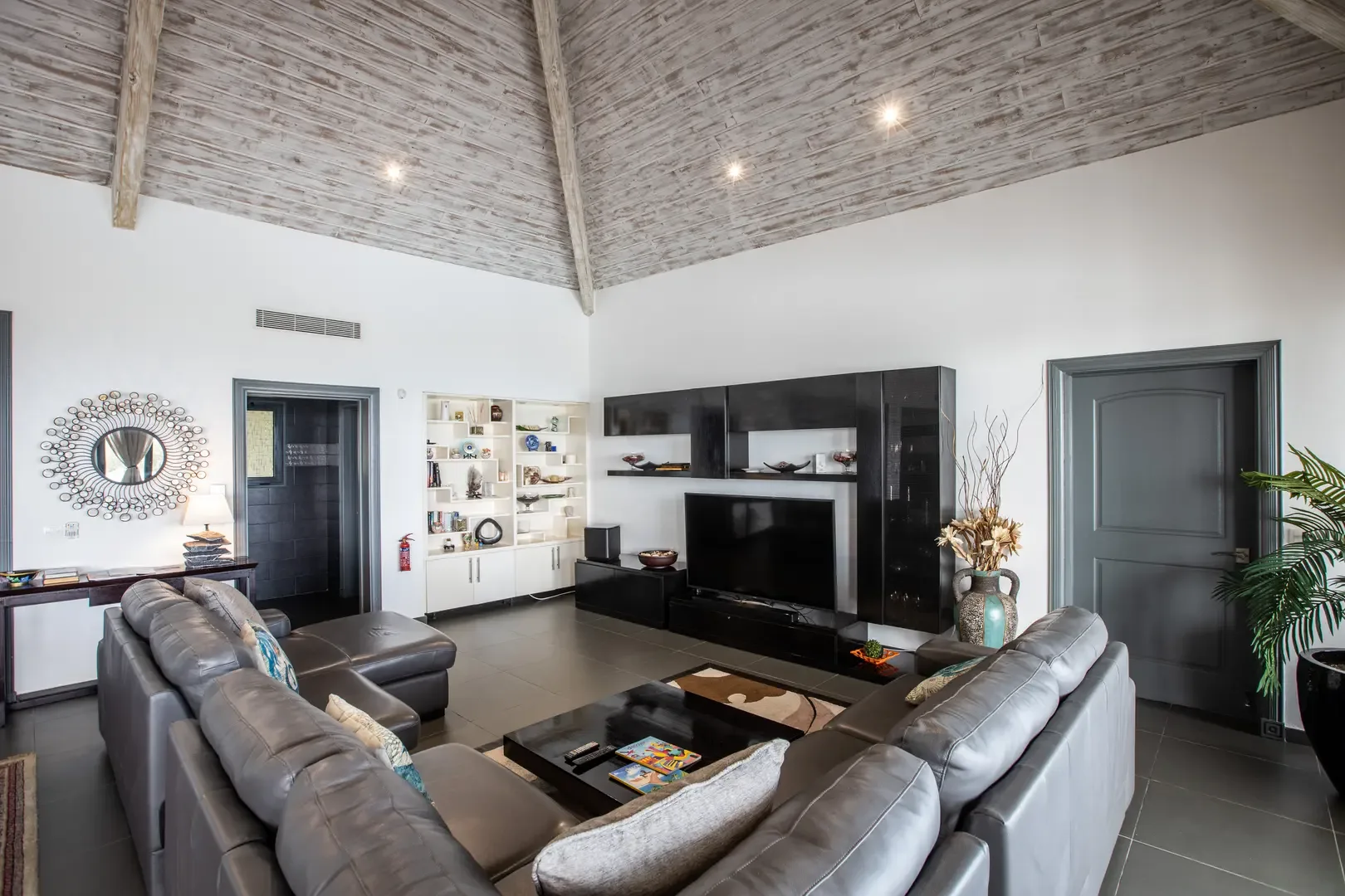 A decorative sunburst mirror anchors the high-ceilinged living area, where a grey leather sectional faces a black entertainment unit and white built-in bookshelves display curated decor.