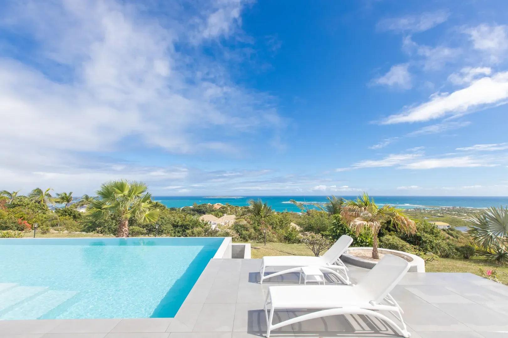 Lounge in style on the modern grey tiled deck as the infinity pool blends seamlessly with the turquoise ocean and lush tropical landscape beyond.