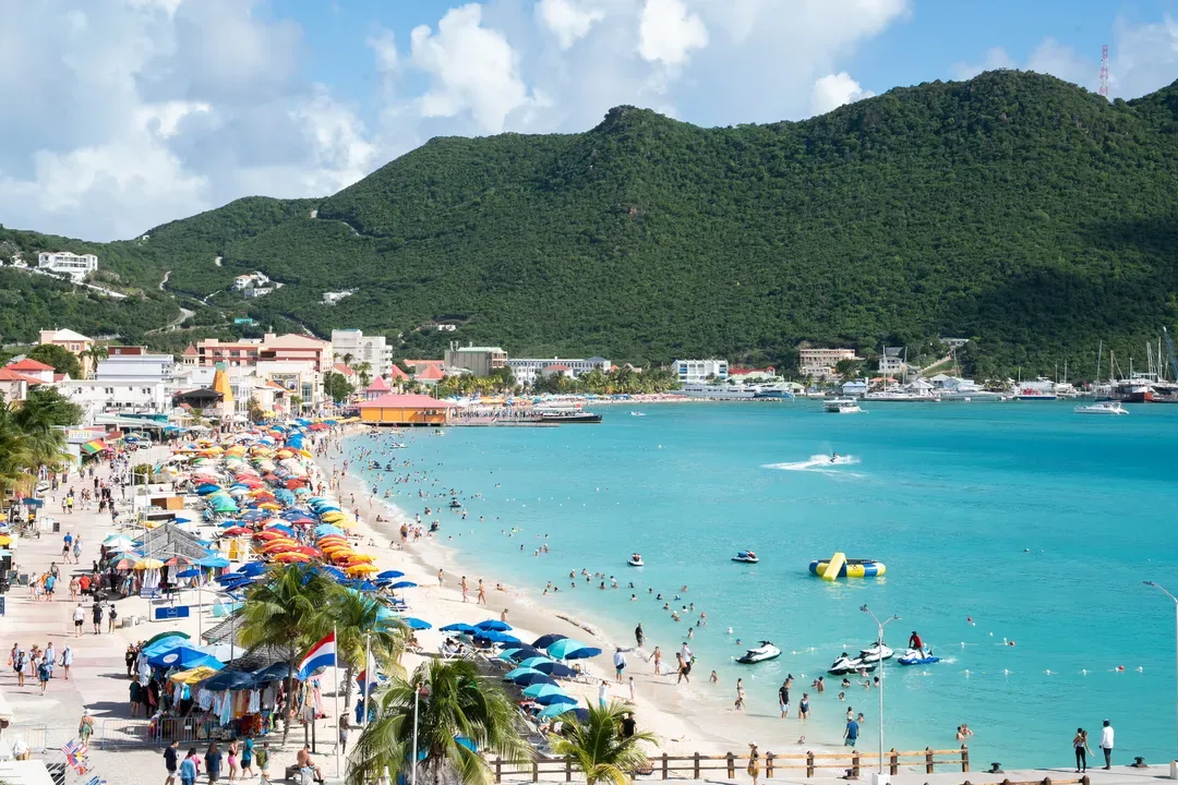 A lively waterfront promenade — colorful beach umbrellas, vendor stalls, and the sparkling turquoise bay inviting you to explore.