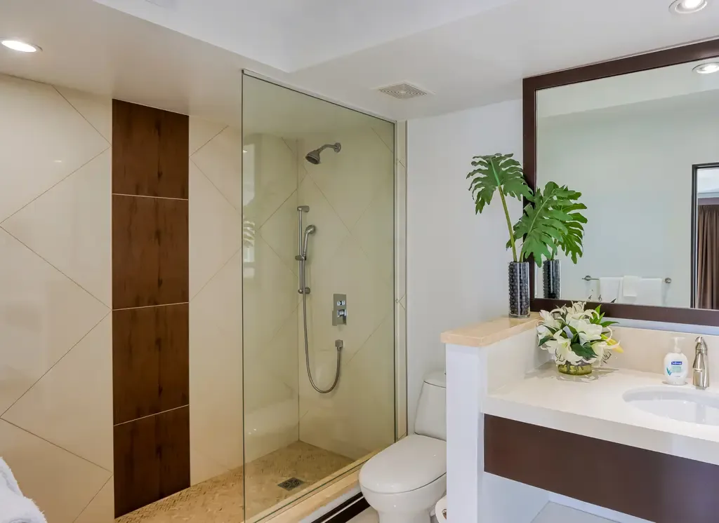 A modern bathroom with glass-enclosed shower featuring a pebble floor, white vanity with large mirror, and decorative plants.