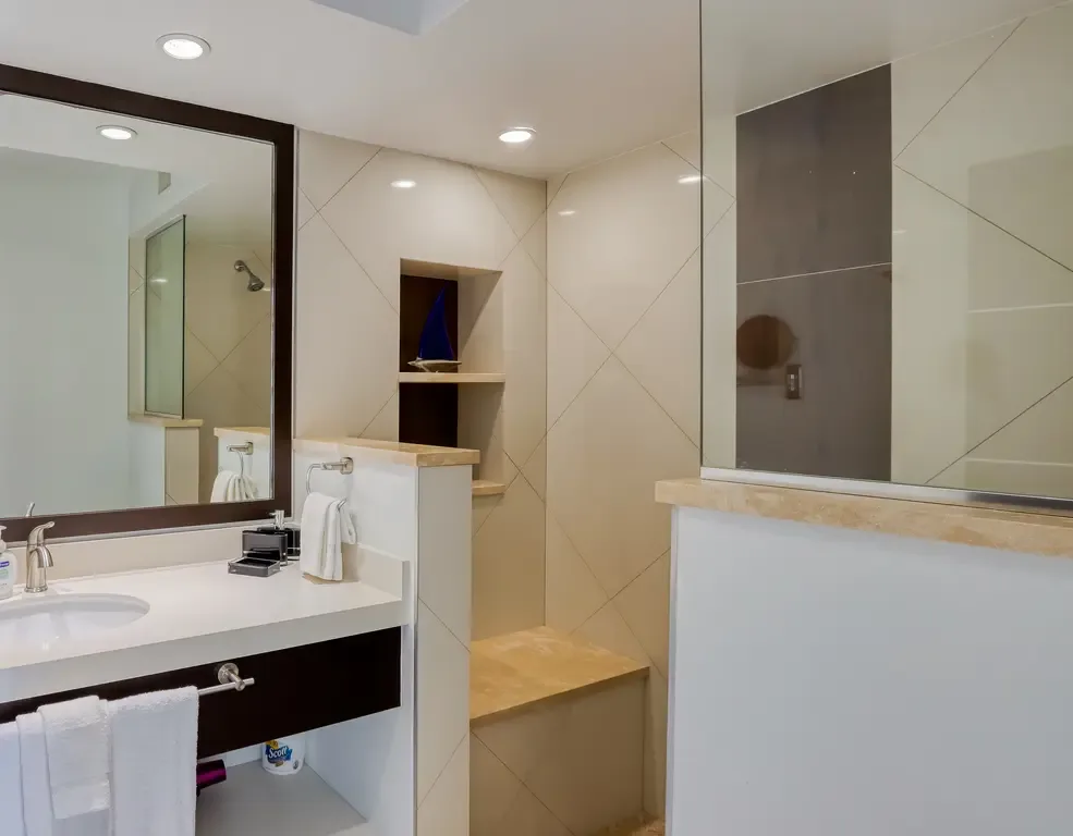 The modern bathroom features a white vanity, dark wood-framed mirror, glass-enclosed shower with built-in bench, and cream-colored tile walls with diamond pattern.