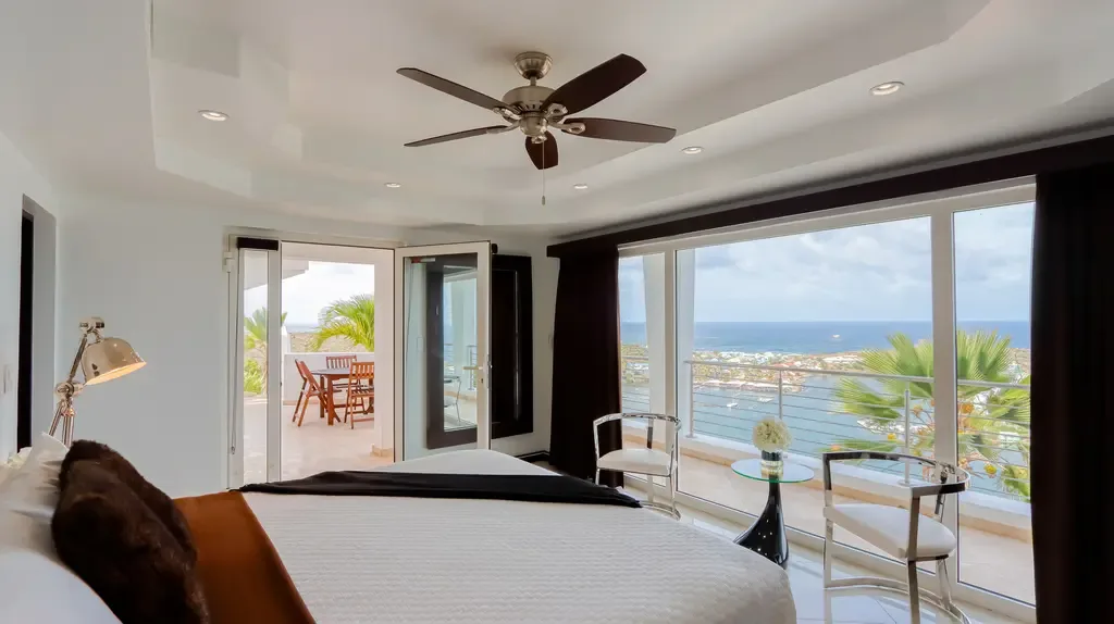 Sliding glass doors open to a private balcony with stunning ocean views and outdoor seating from this bedroom with ceiling fan.