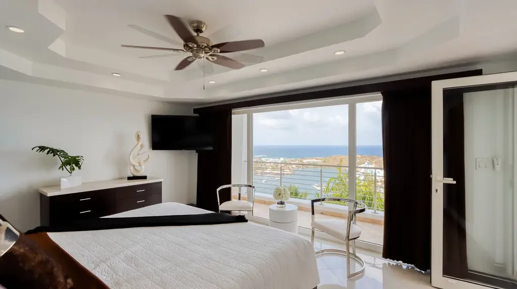 Large glass doors open to a balcony with ocean and coastal views from this modern bedroom with a dark wood dresser and coffered ceiling with fan.