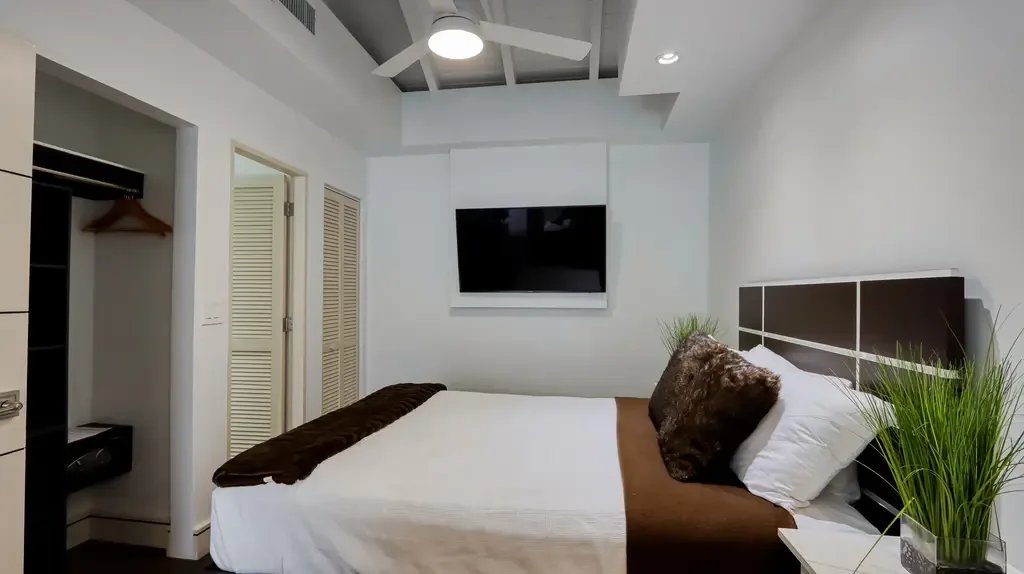 A contemporary bedroom features a white bed with brown accents, wall-mounted TV, ceiling fan, and louvered doors.