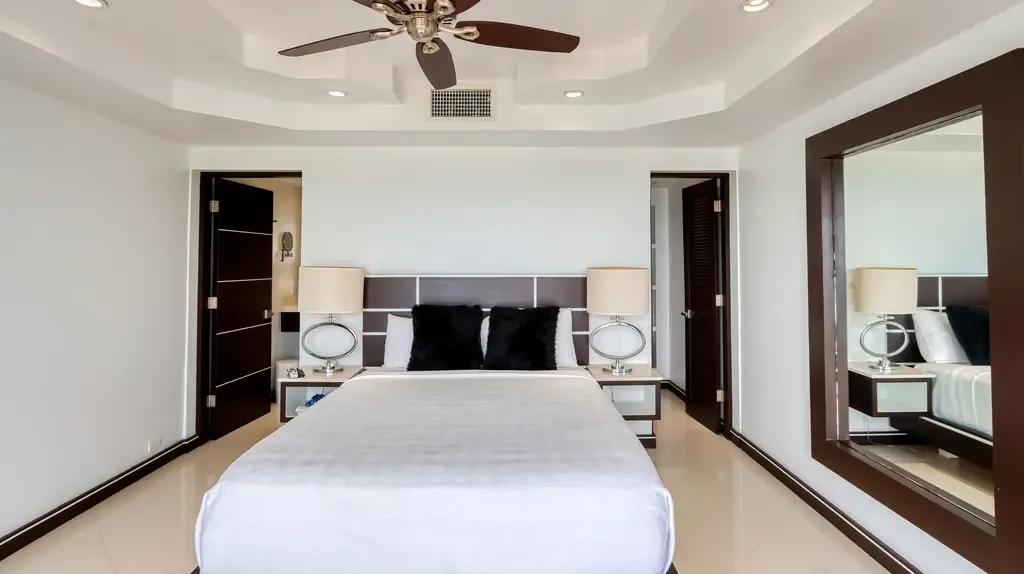 A modern bedroom features a dark paneled headboard, black accent pillows, two nightstands with large circular lamps, and a large dark-framed floor mirror.