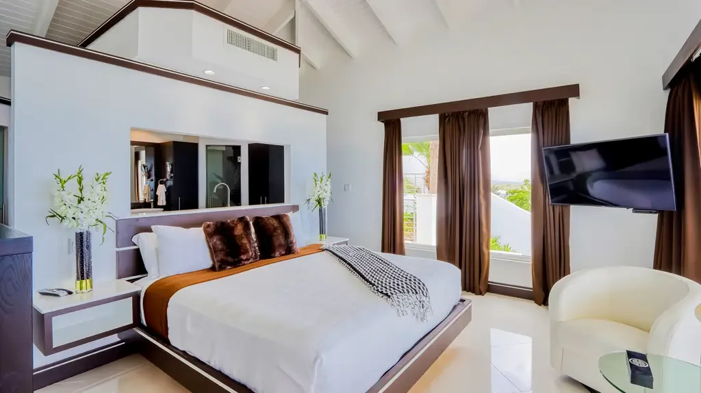 The modern master suite boasts vaulted ceilings, a king bed with plush brown accents, white furnishings, and a view into the ensuite bathroom.