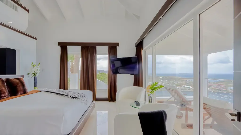 Large glass sliding doors reveal an expansive ocean view from this modern white bedroom with a wall-mounted TV and white armchair.