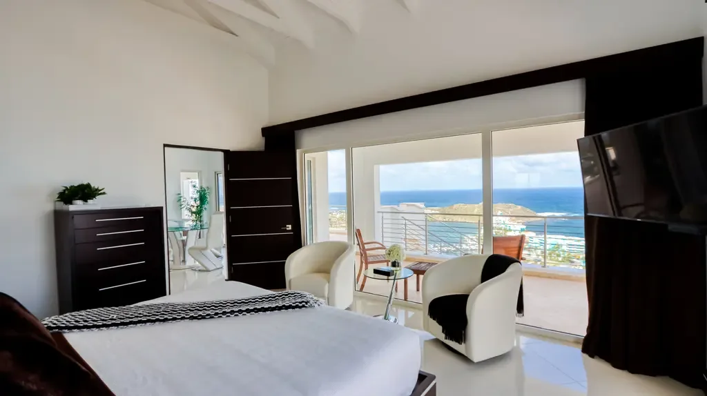 Expansive glass doors open to a terrace with panoramic ocean and coastline views from this luxurious bedroom with white bucket-style armchairs.