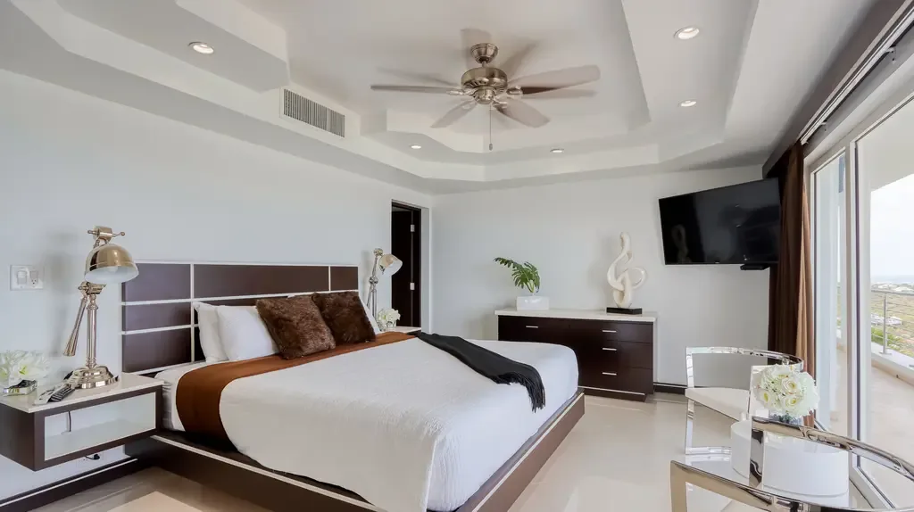 A modern master bedroom features a large bed with plush brown accent pillows, dark wood furniture, and sliding glass doors to a balcony with scenic views.