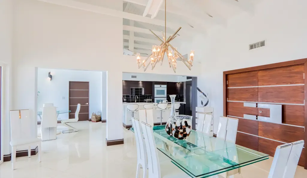 The open-concept dining space features a sculptural gold chandelier, glass-top table with high-back white chairs, and seamless connection to the modern kitchen.