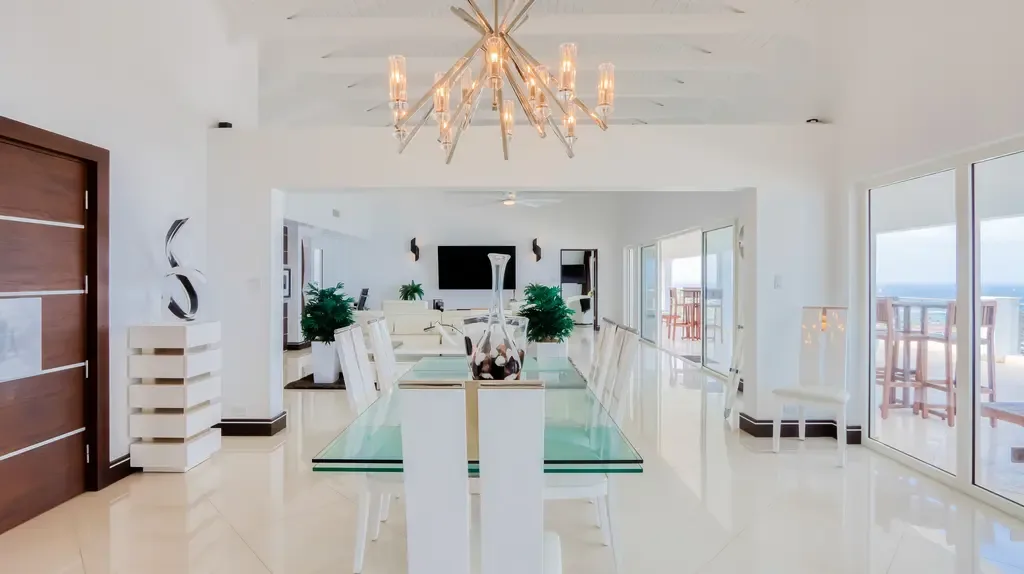 A bright open-plan space showcases a glass-top dining table, modern chandelier, and seamless connection to the living area with ocean views through glass doors.