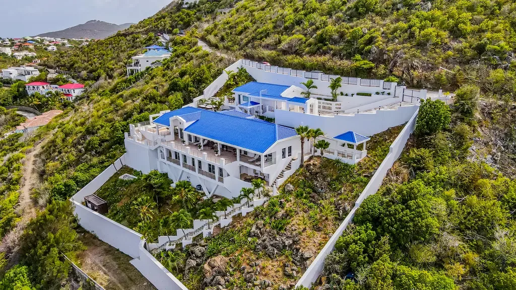 An elevated aerial view reveals this stunning white villa with striking blue roofs, multiple levels, and lush tropical landscaping on an exclusive Oyster Pond hillside.