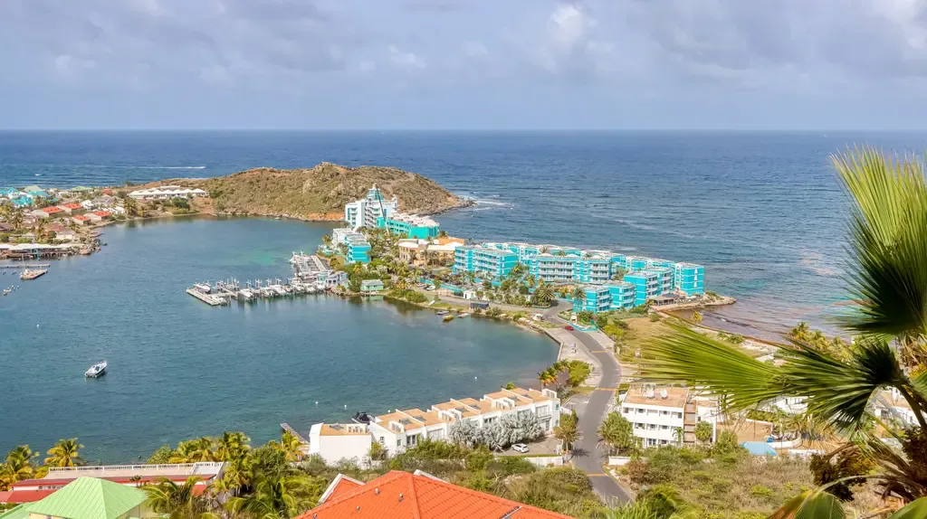 A wide aerial shot showcases the coastal landscape with turquoise resort buildings, a marina with boats, and a peninsula extending into the ocean.