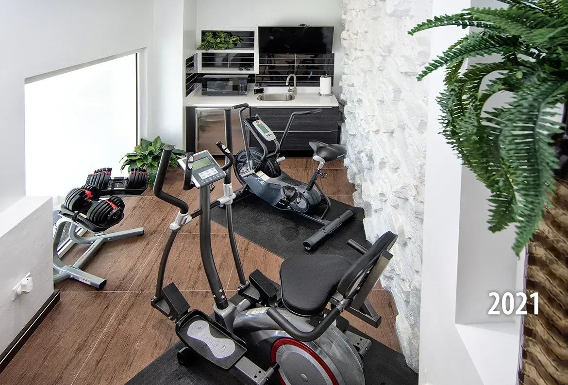 The modern home fitness center includes an elliptical machine, stationary bike, adjustable dumbbells, and a small kitchenette with textured stone wall accents.