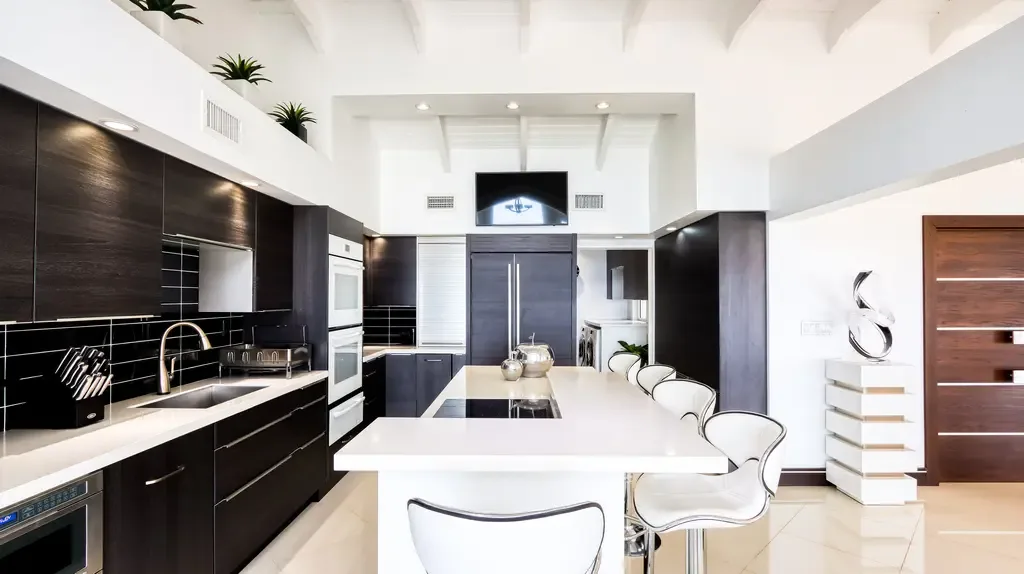 The modern high-end kitchen features dark wood cabinetry, white countertops, a large island with integrated seating, and professional-grade appliances beneath vaulted beamed ceilings.
