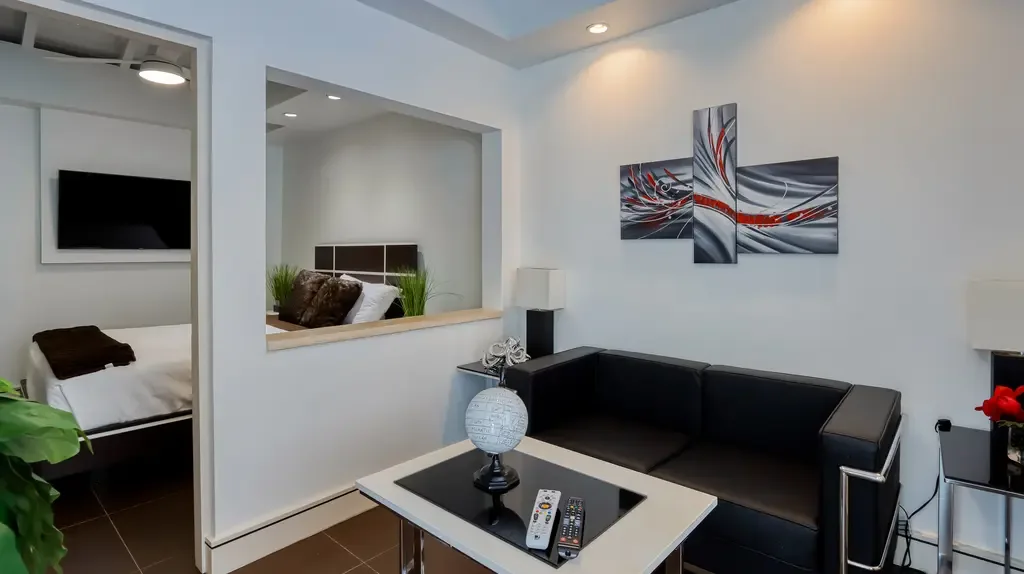 A black leather sofa, coffee table with decorative globe, and modern abstract wall art connect to an adjacent bedroom area with wall-mounted TV.