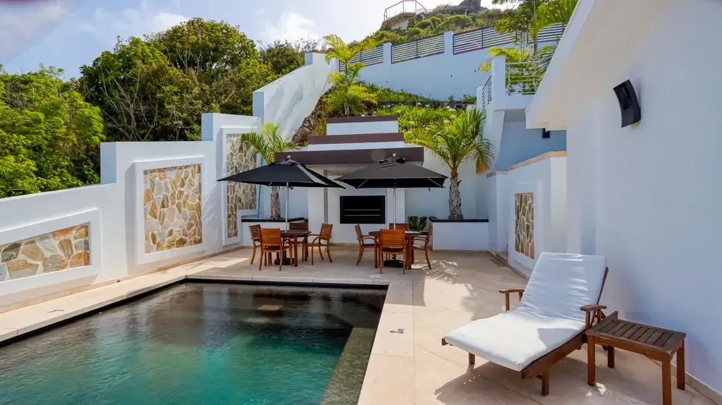 The outdoor pool area features stone-inlaid privacy walls, two dining sets with black umbrellas, and a built-in outdoor grill area.