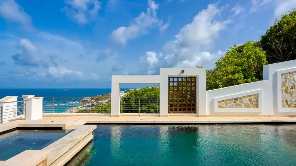 The elevated infinity-edge pool area features a white geometric privacy wall with stone accents and panoramic ocean views over a coastal town.
