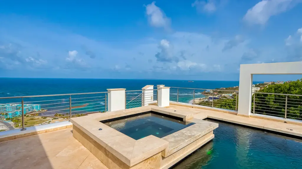 A luxurious rooftop terrace showcases a stone-clad hot tub spilling into an infinity-edge pool with unobstructed panoramic views of the turquoise ocean and coastline.