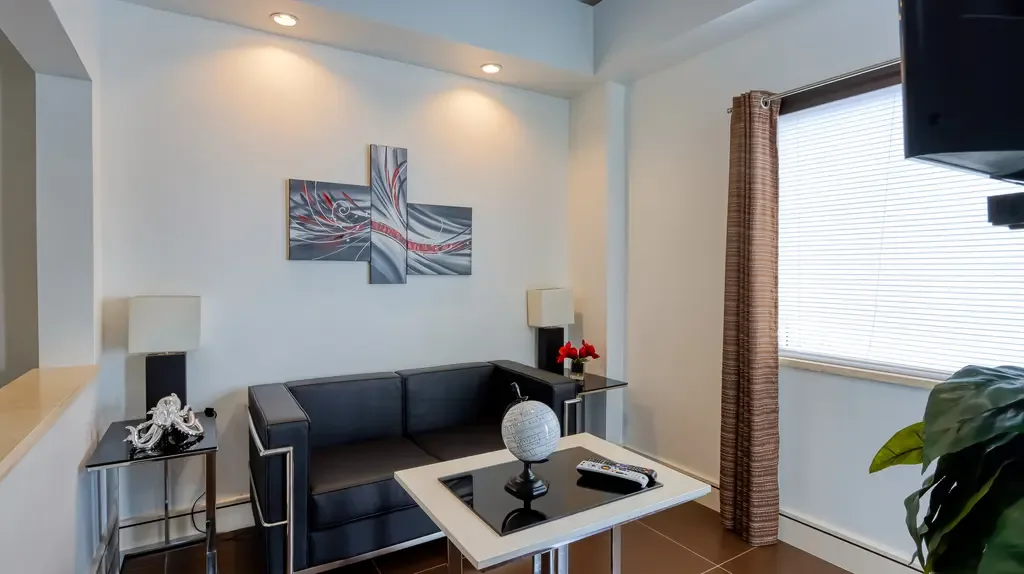 A compact modern sitting area features a black leather-style loveseat, white coffee table with globe, side tables with lamps, and abstract wall art.