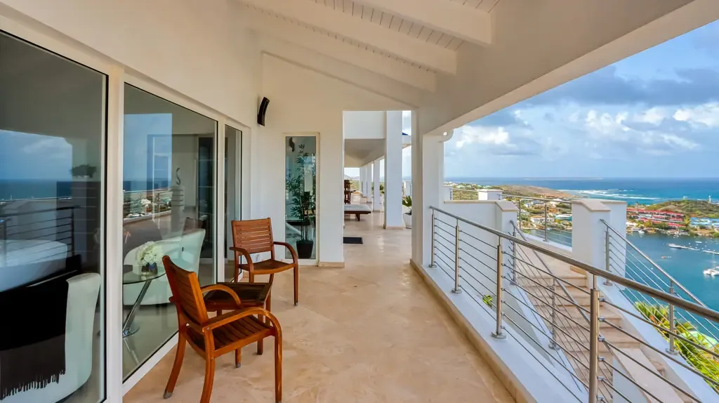 A covered balcony with wooden chairs and modern cable railings offers spectacular panoramic views of the bay and ocean.