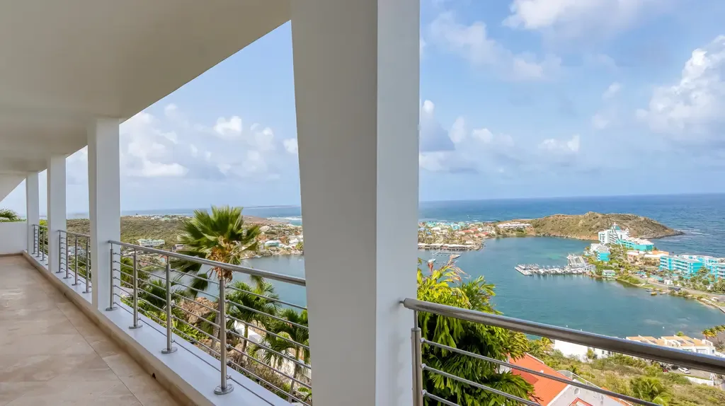 A long spacious white-tiled terrace with modern metal railings offers breathtaking panoramic views of a tropical bay, coastal town, and ocean.