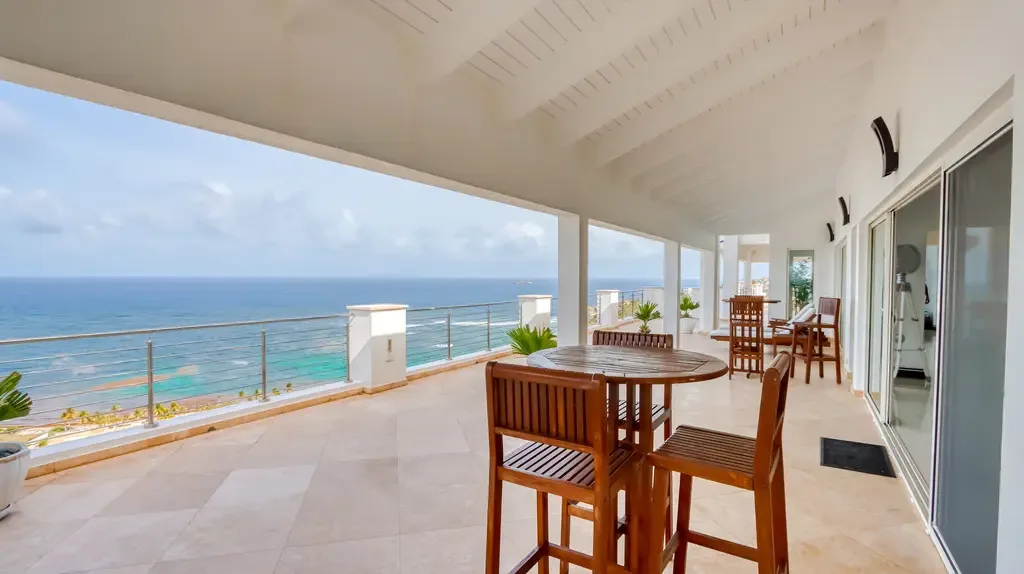 Wooden high-top seating and lounge furniture frame expansive turquoise ocean views through sleek cable railings, with white architecture and vaulted ceilings.