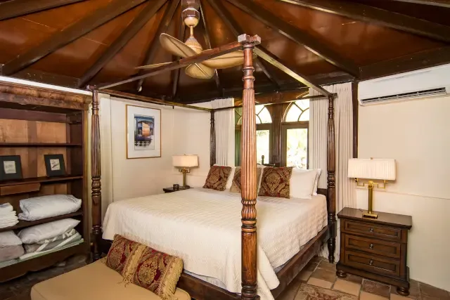 Your romantic four-poster bed awaits — white linens and arched windows frame your island dreams.