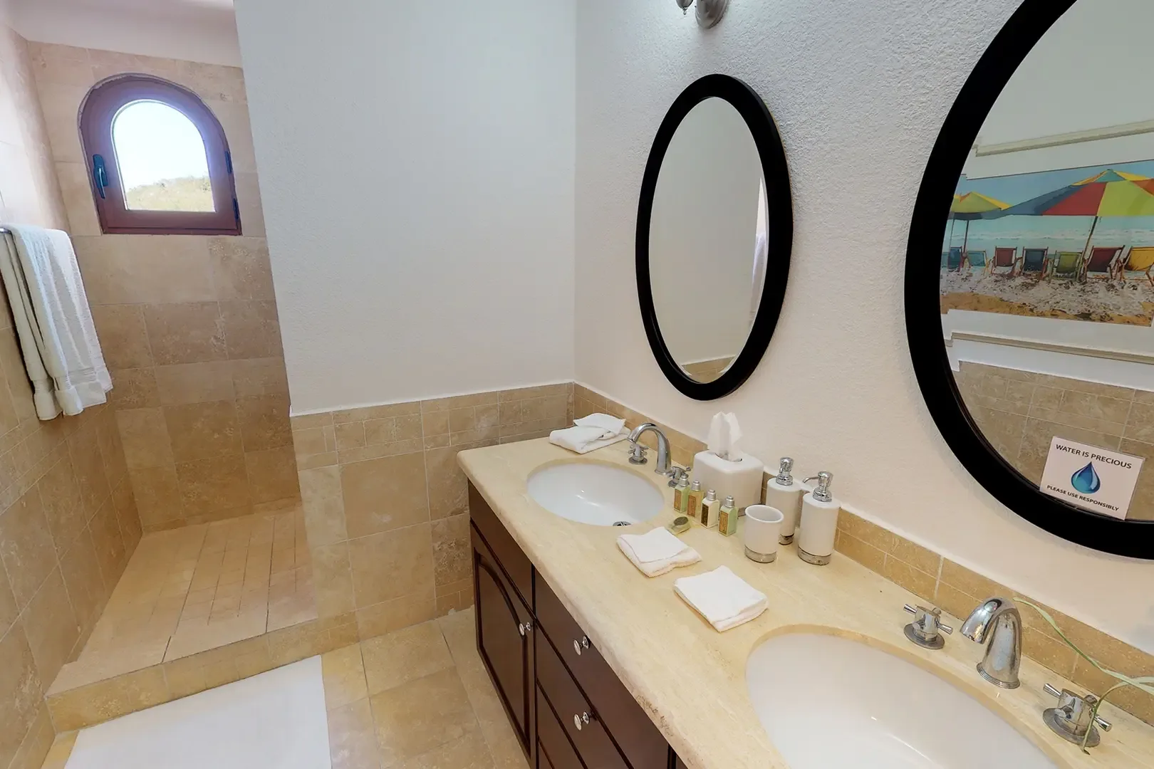 Double vanity with stone countertops and oval mirrors, walk-in shower with natural tile, and decorative arched window.