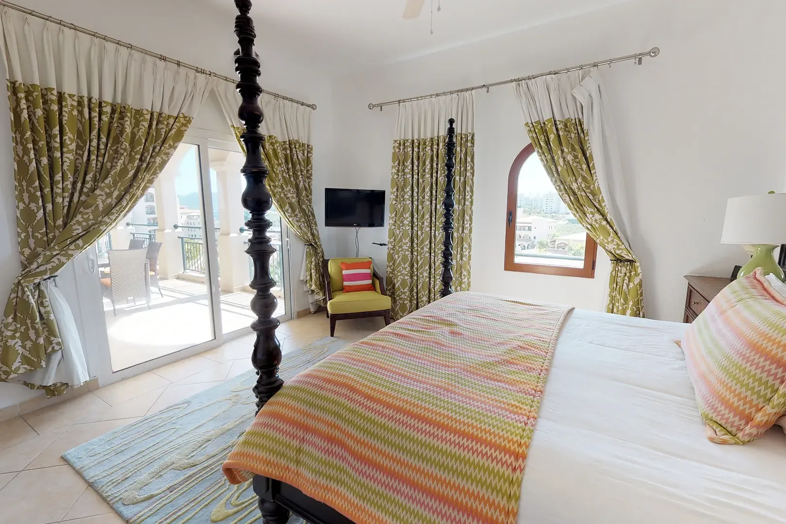 A dark wood four-poster bed with colorful chevron-patterned throw anchors this bedroom, with large glass sliding doors to the balcony framed by patterned green and white curtains.