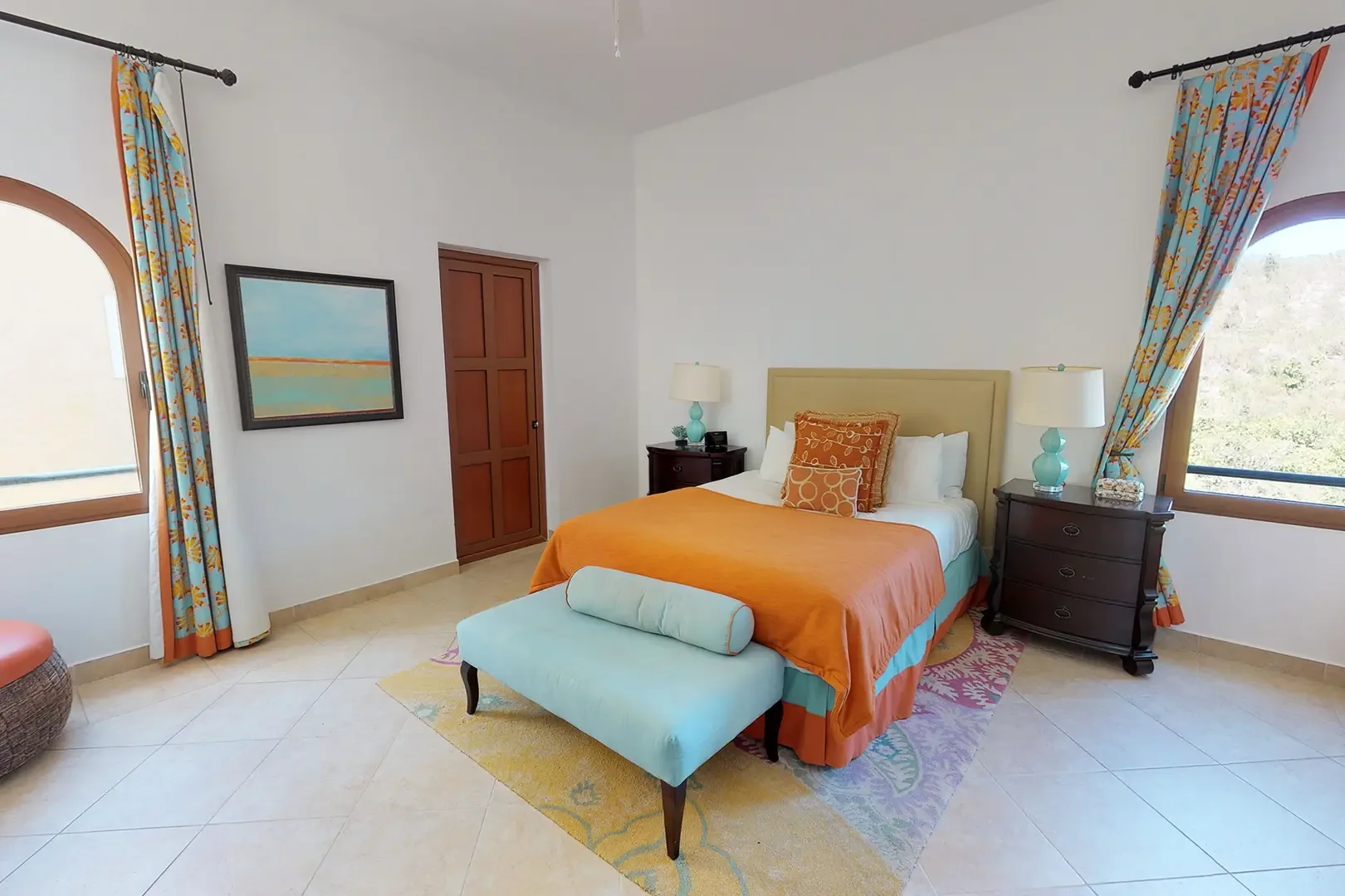 A well-lit guest bedroom with queen bed features orange and teal bedding, matching bedside lamps, light blue bench, patterned curtains, and arched windows.