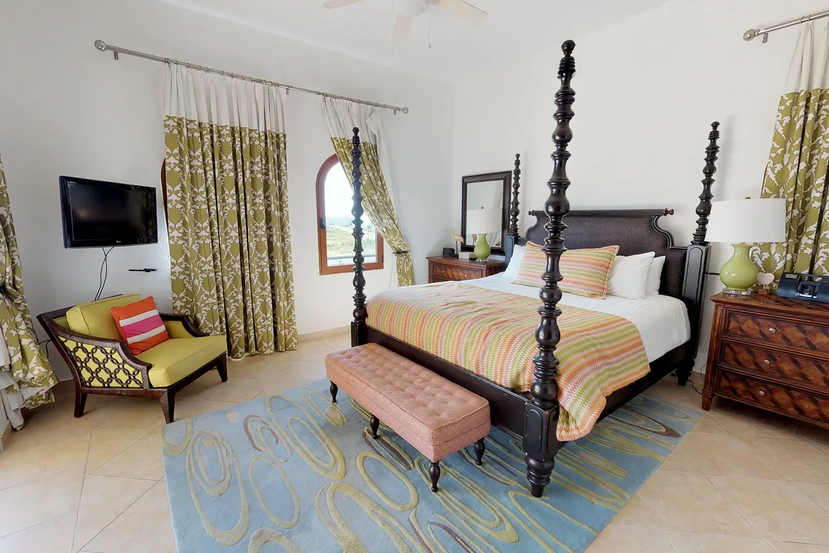 The spacious master bedroom features a dark wood four-poster bed with colorful patterned bedspread, yellow accent chair, light blue patterned rug, and olive green and white curtains.