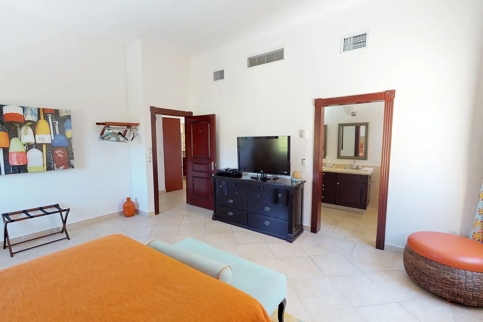 King bed with orange bedding, dark wood dresser with flat-screen TV, and nautical-themed wall art, with bathroom visible through open doorway.