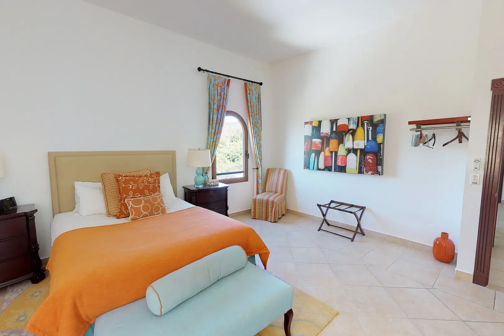 Bright bedroom with orange throw and light blue bench, striped accent chair, colorful buoy art piece, and patterned curtains framing an arched window.