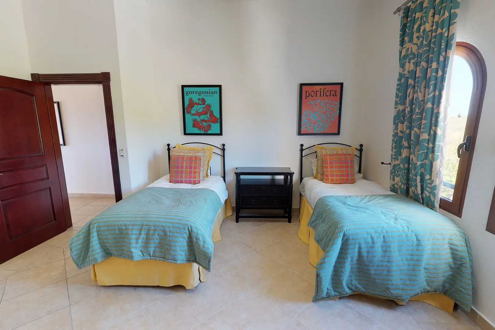 Twin bedroom with teal and yellow bedding, botanical wall art, dark wood nightstand, and patterned curtains.