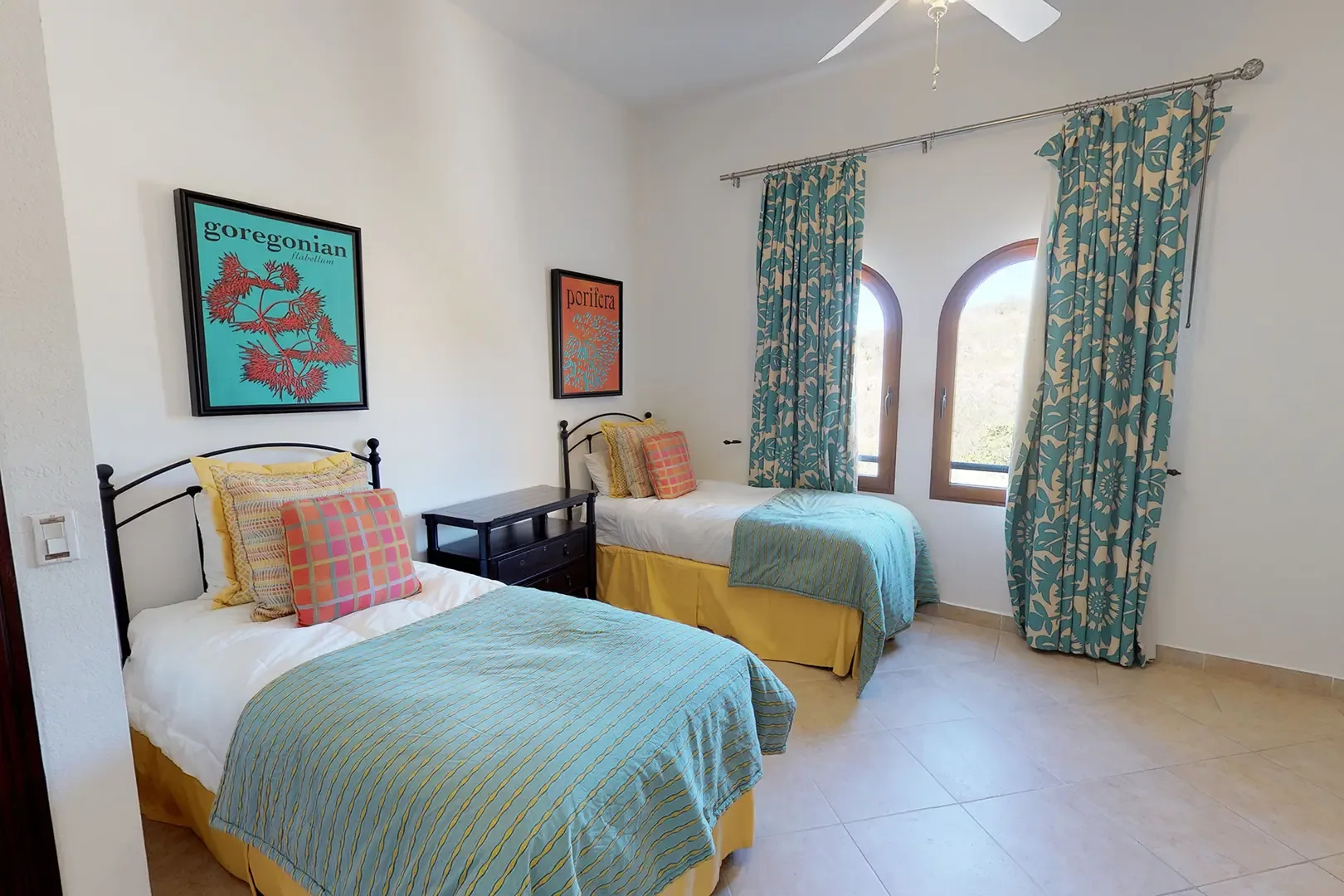 Two twin beds with vibrant bedding, arched windows with patterned curtains, and botanical-themed wall art.