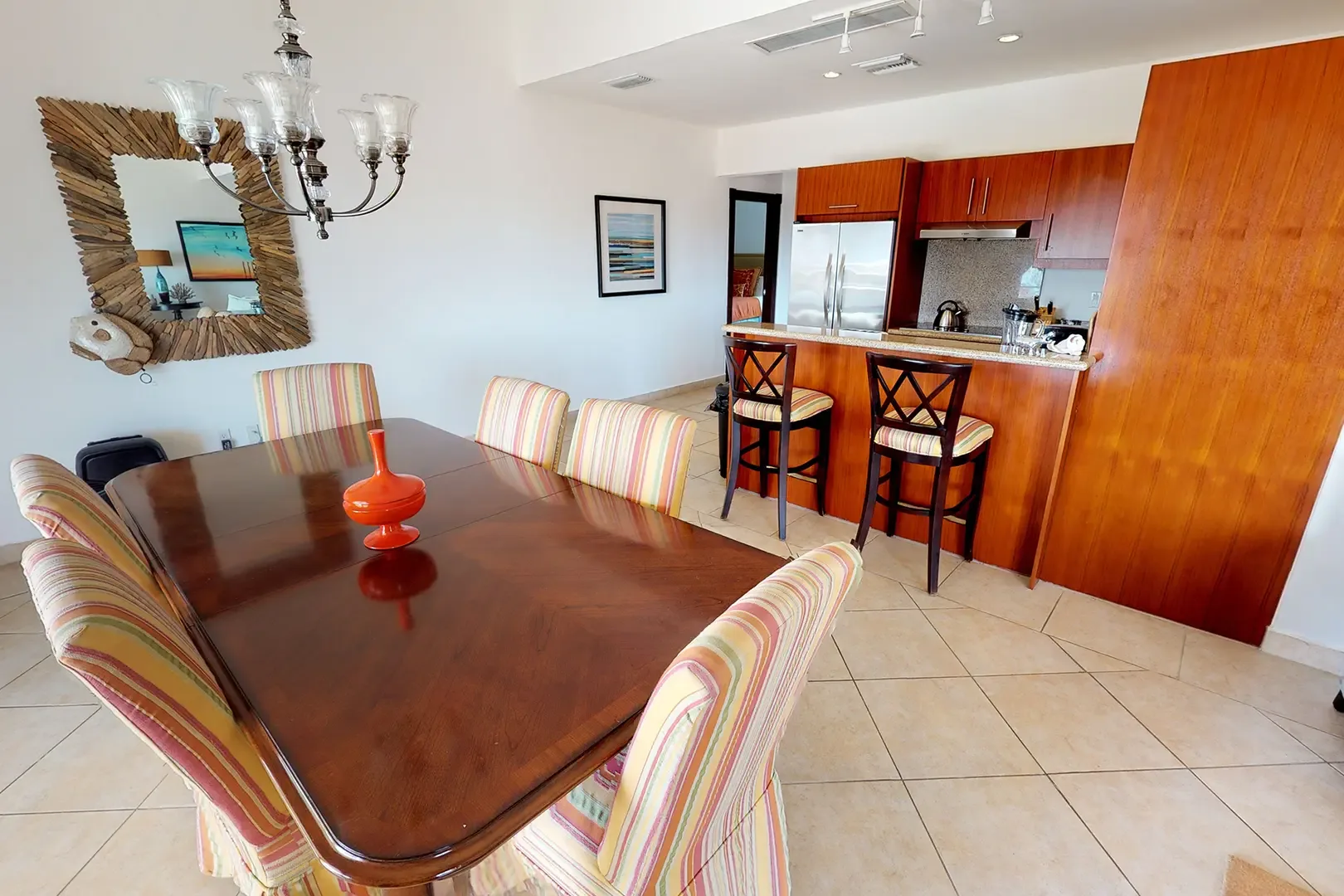 A large polished wood dining table with six striped upholstered chairs and decorative orange vase sits adjacent to the kitchen with breakfast bar and stainless steel appliances.