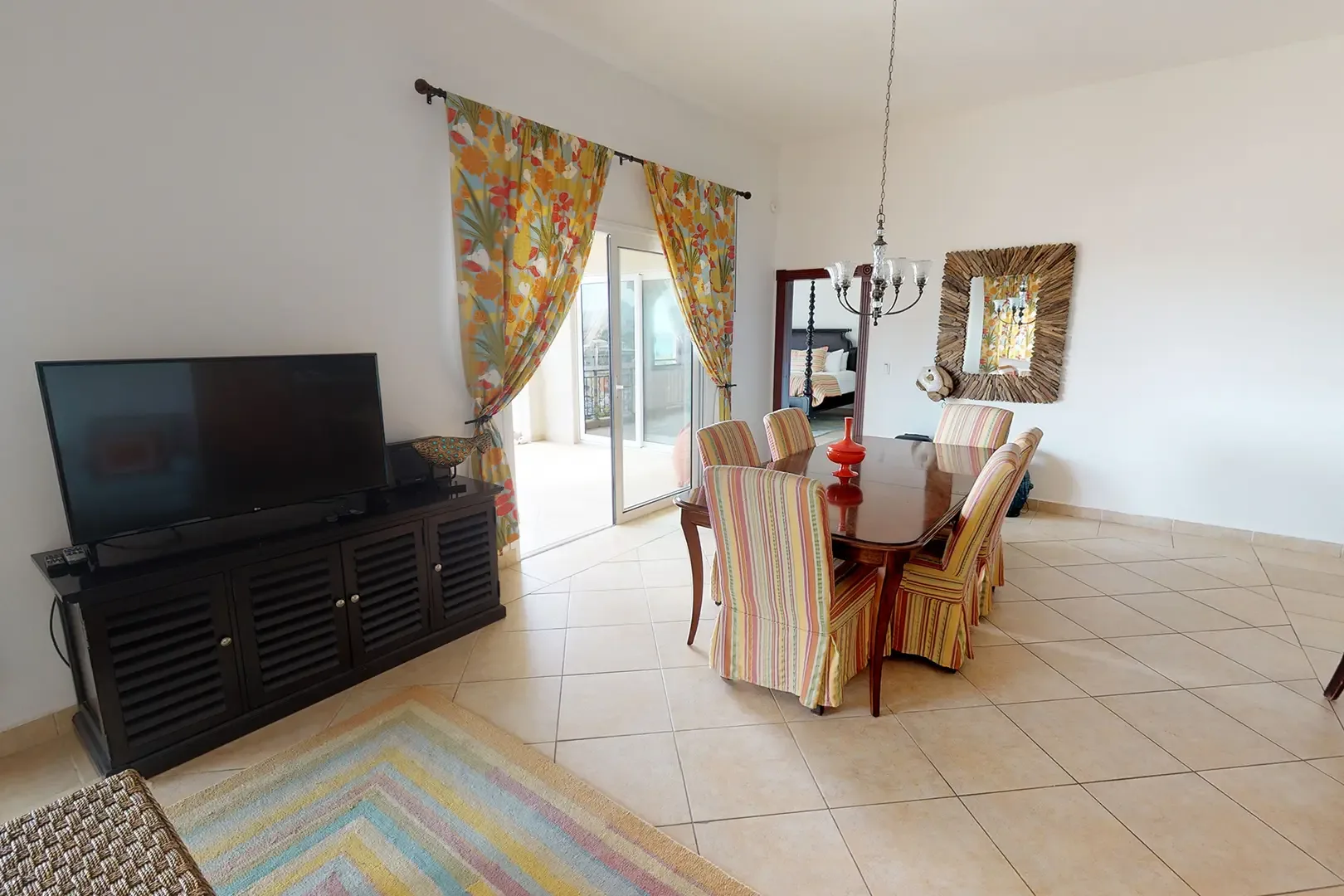 The open-concept space features a dark wood dining set with six striped chairs, large media console with flat-screen TV, vibrant tropical curtains, and driftwood-framed mirror with crystal chandelier.