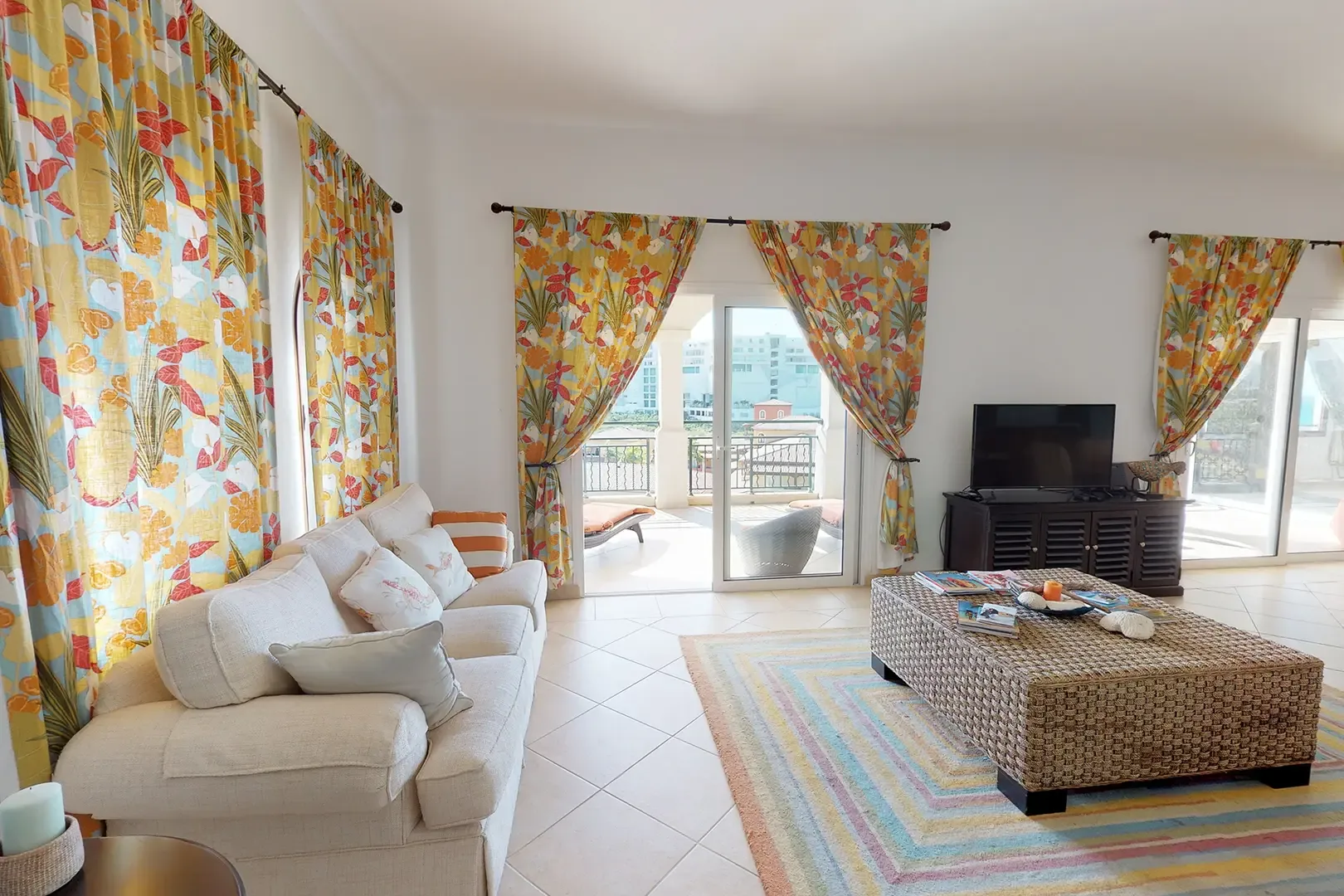 A bright living area features floral curtains, cream-colored sofa with accent pillows, large woven coffee table, and colorful striped rug with sliding glass doors to the balcony.