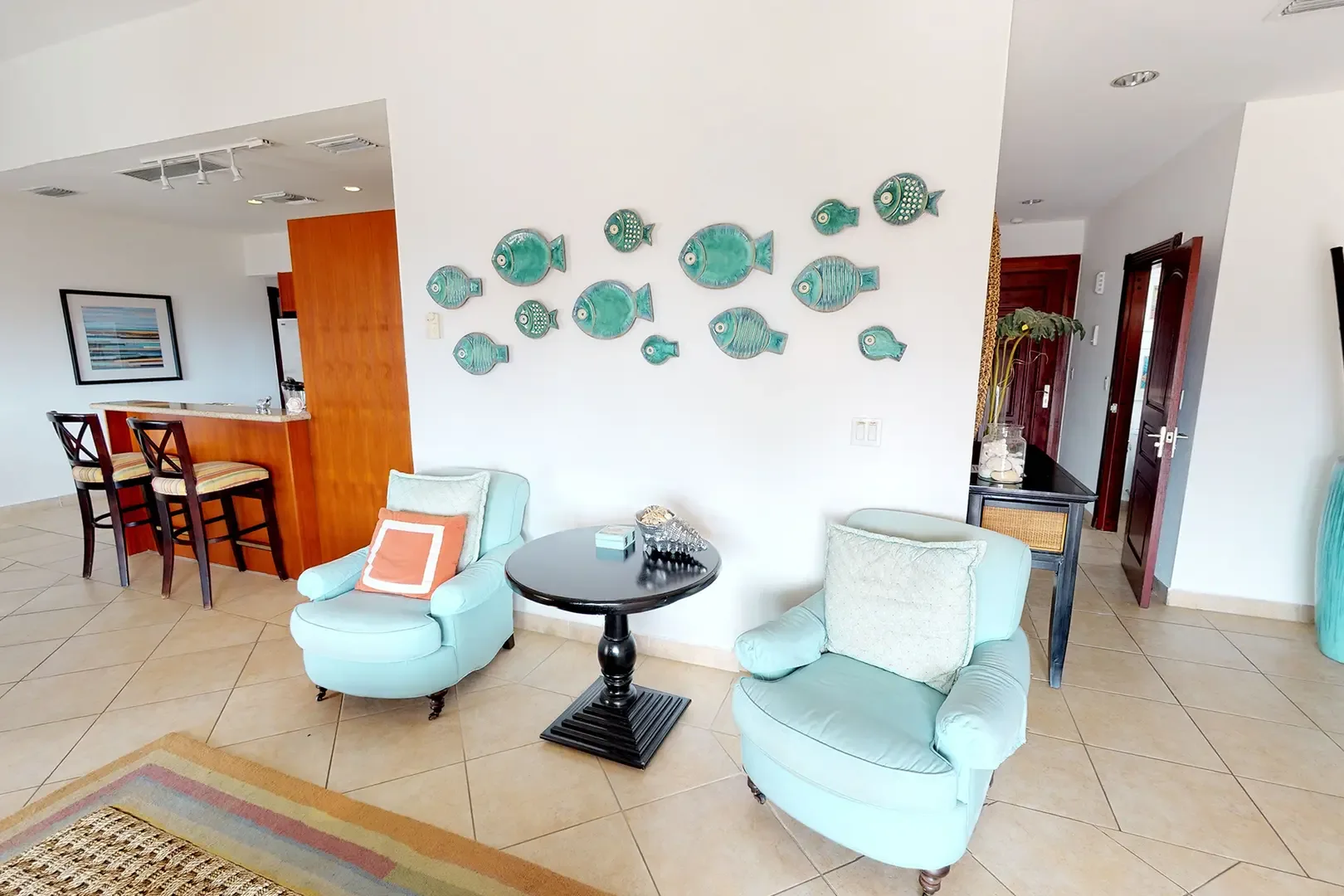 Two light blue armchairs and a black pedestal side table sit beneath a wall decorated with a school of ceramic fish, with breakfast bar and kitchen in the background.