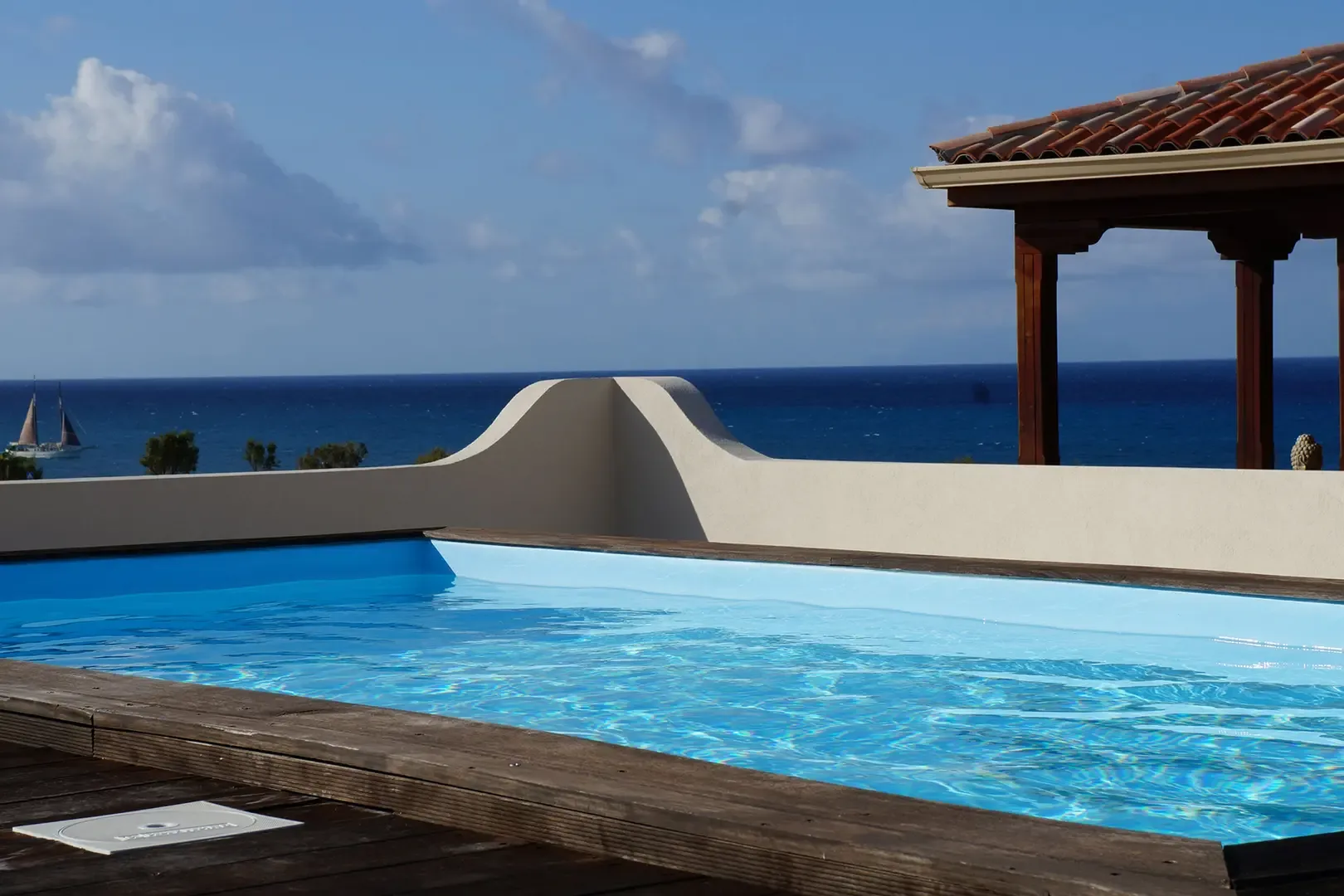 The infinity-style pool with crystal-clear water and wooden decking frames a stunning panoramic ocean view with sailboats in the distance.